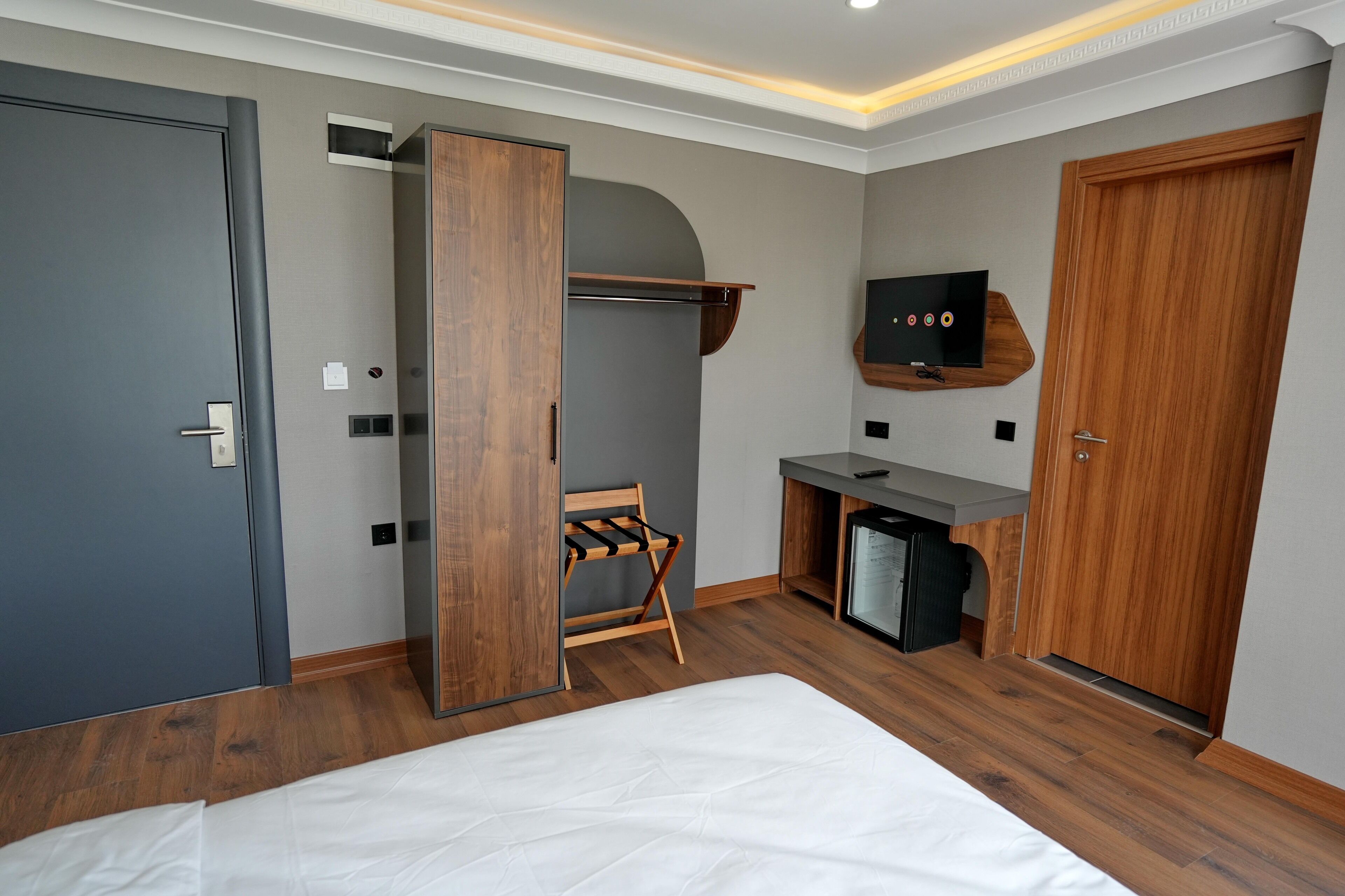 A modern hotel room with a bed, a wooden wardrobe, a desk with a TV, and a small seating area.