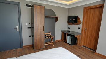 A modern hotel room with a bed, a wooden wardrobe, a desk with a TV, and a small seating area.