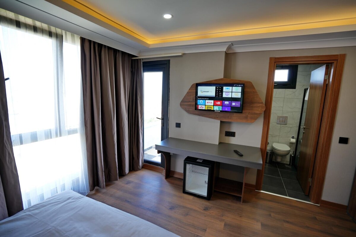 A hotel room with a bed, a desk with a flat-screen TV, a small fridge, and a bathroom visible through an open door.