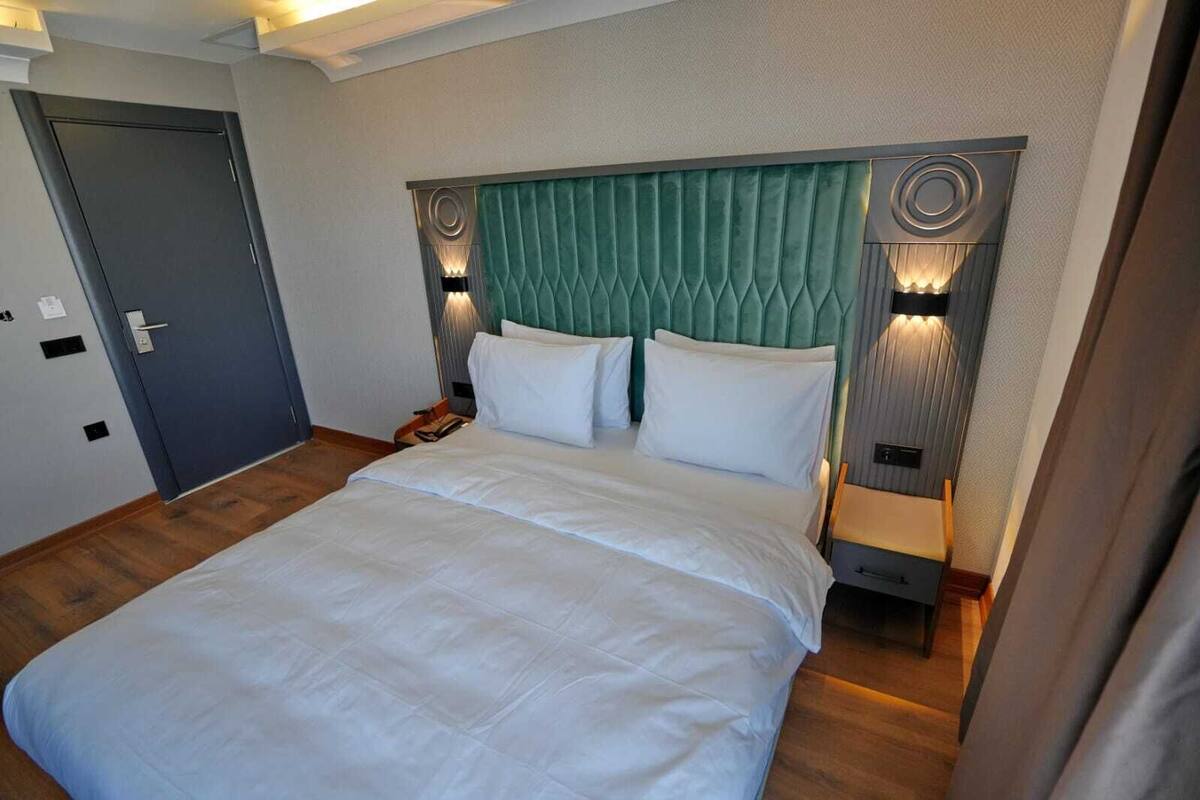 Une chambre d’hôtel avec un grand lit, une tête de lit recouverte d’un tissu vert, des lampes de chevet et une porte.