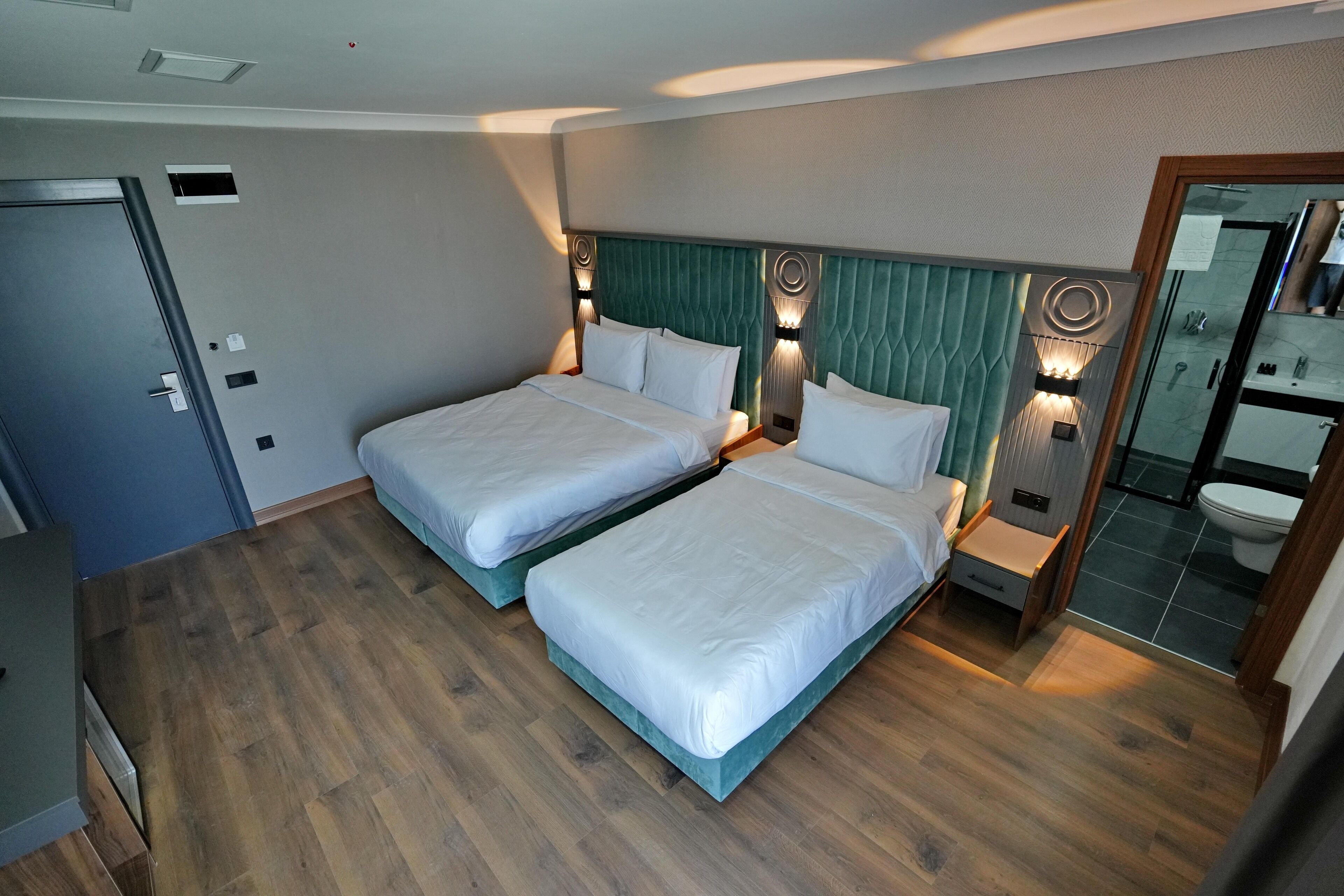 A hotel room with two beds, a wooden floor, a green headboard, and a bathroom with a glass shower.