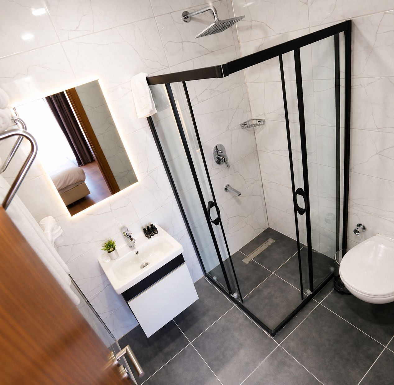 A modern bathroom with a glass-enclosed shower, a white sink with toiletries, and a mirror reflecting a bedroom.