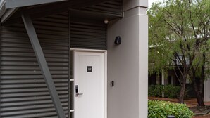 A modern building entrance with a white door marked with the number 112, a metal ramp, and a small garden area with a tree and a potted plant. - At Forest Stay | Apartment in Margaret River | New Listing (Margaret River)