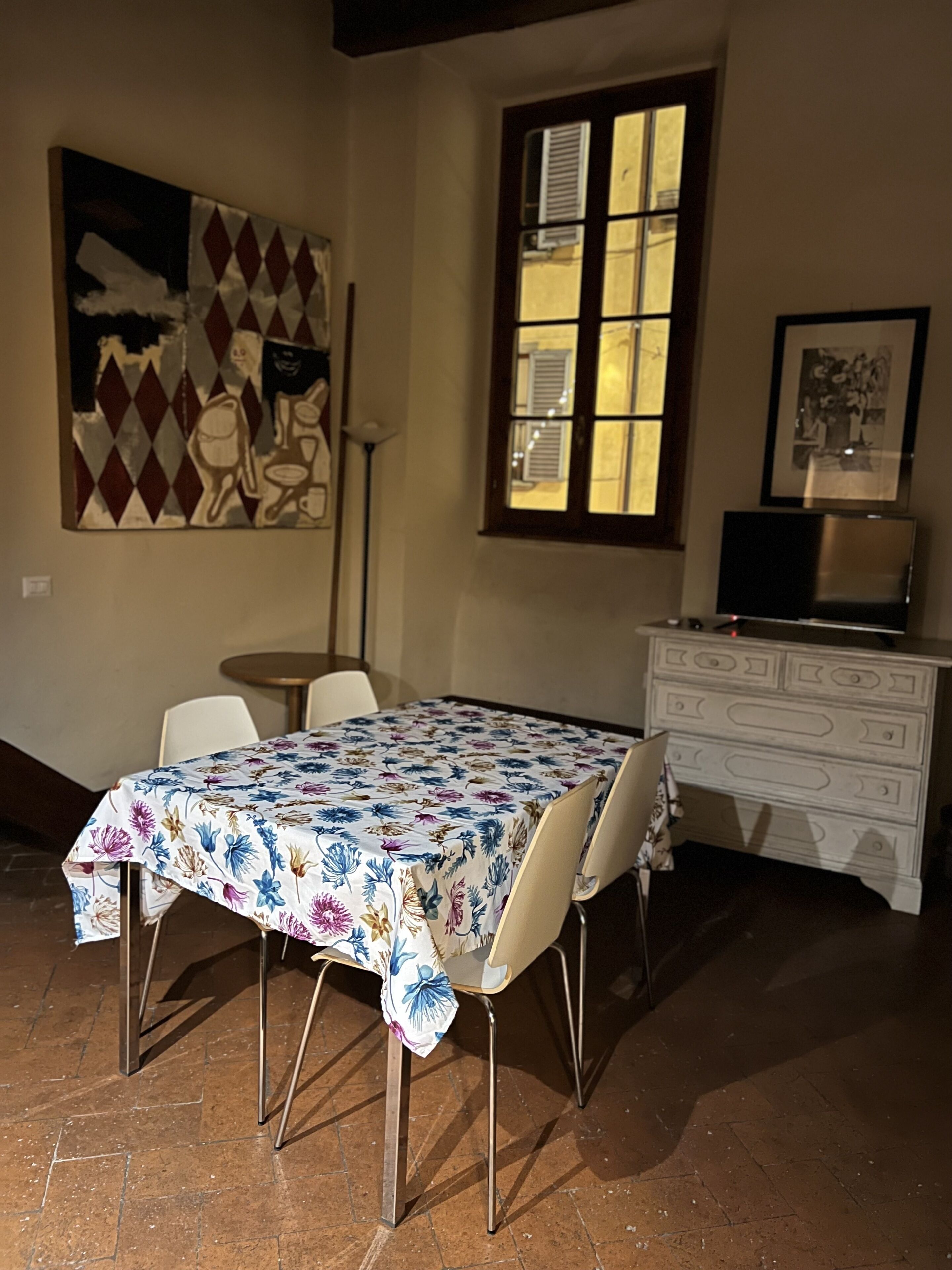 A dining area with a table covered in a floral tablecloth, chairs, a dresser, a painting on the wall, and a window with shutters.