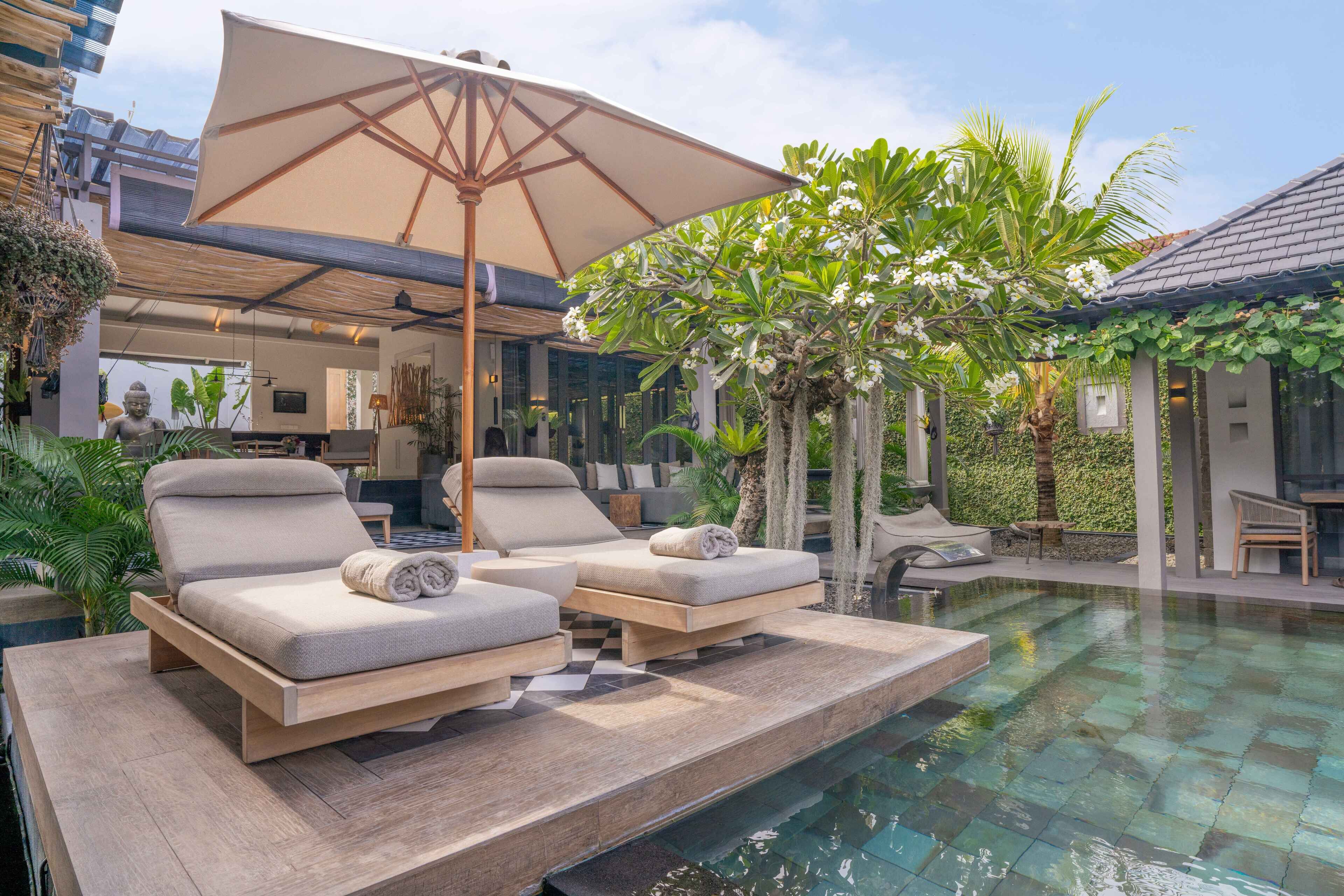 A poolside area with lounge chairs and a large umbrella, surrounded by greenery and a modern building.