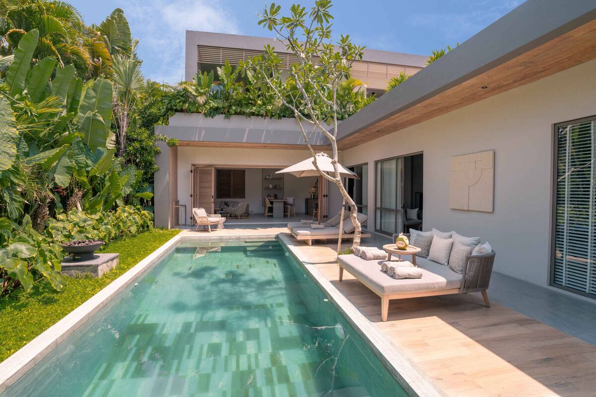 A modern pool area with a built-in seating area, surrounded by lush greenery and a contemporary house.