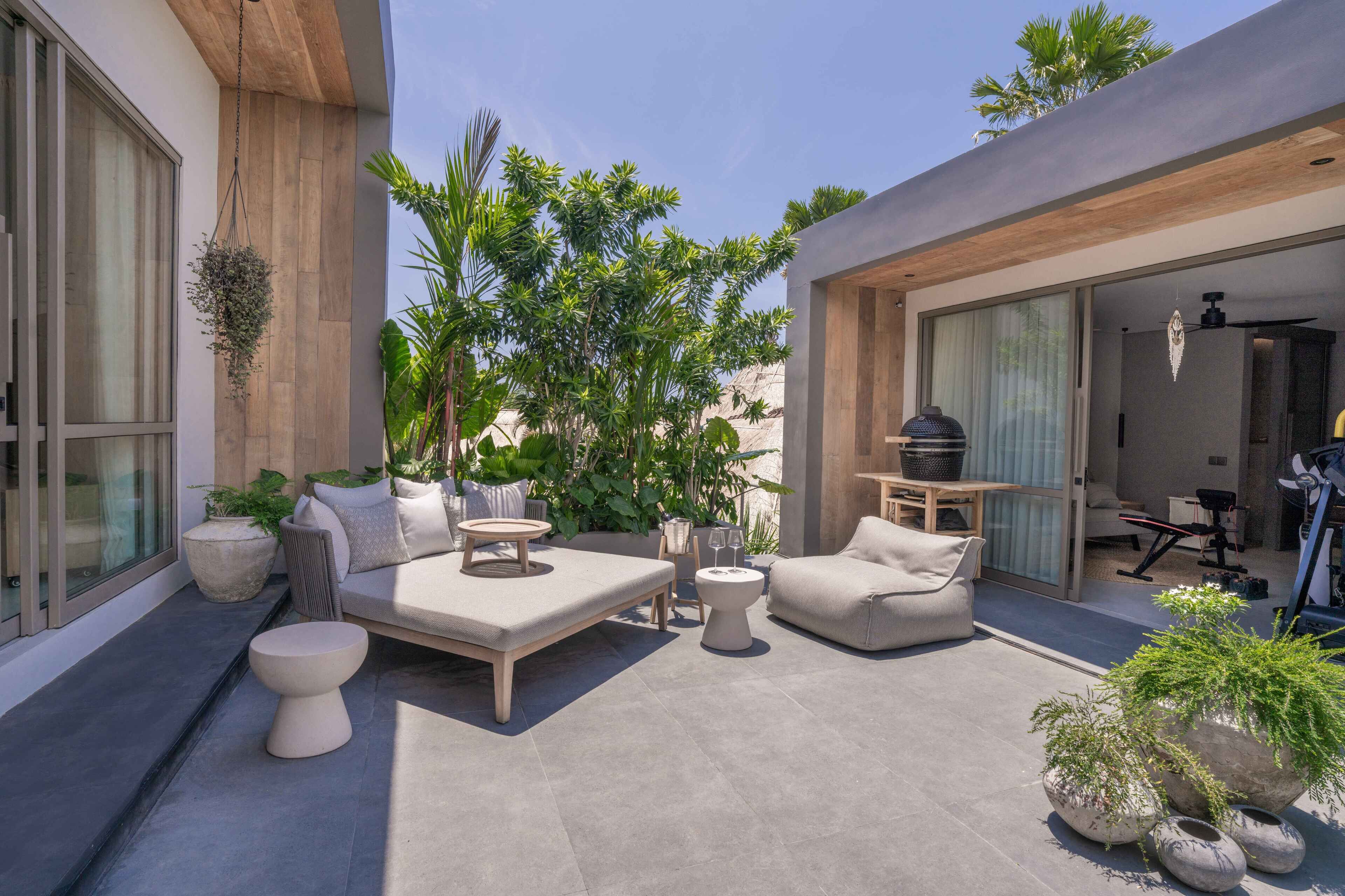 Un espacio exterior moderno para el descanso con sofá, mesa de centro y zona de estar, rodeado de plantas en macetas y un ventanal grande.