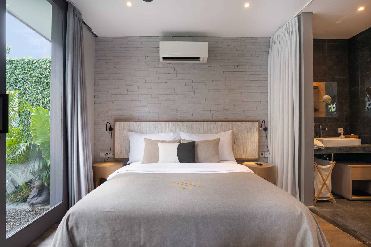 A modern hotel room with a brick wall, a large bed with a grey bedspread, a bedside table with a lamp, and a view of greenery through a window.