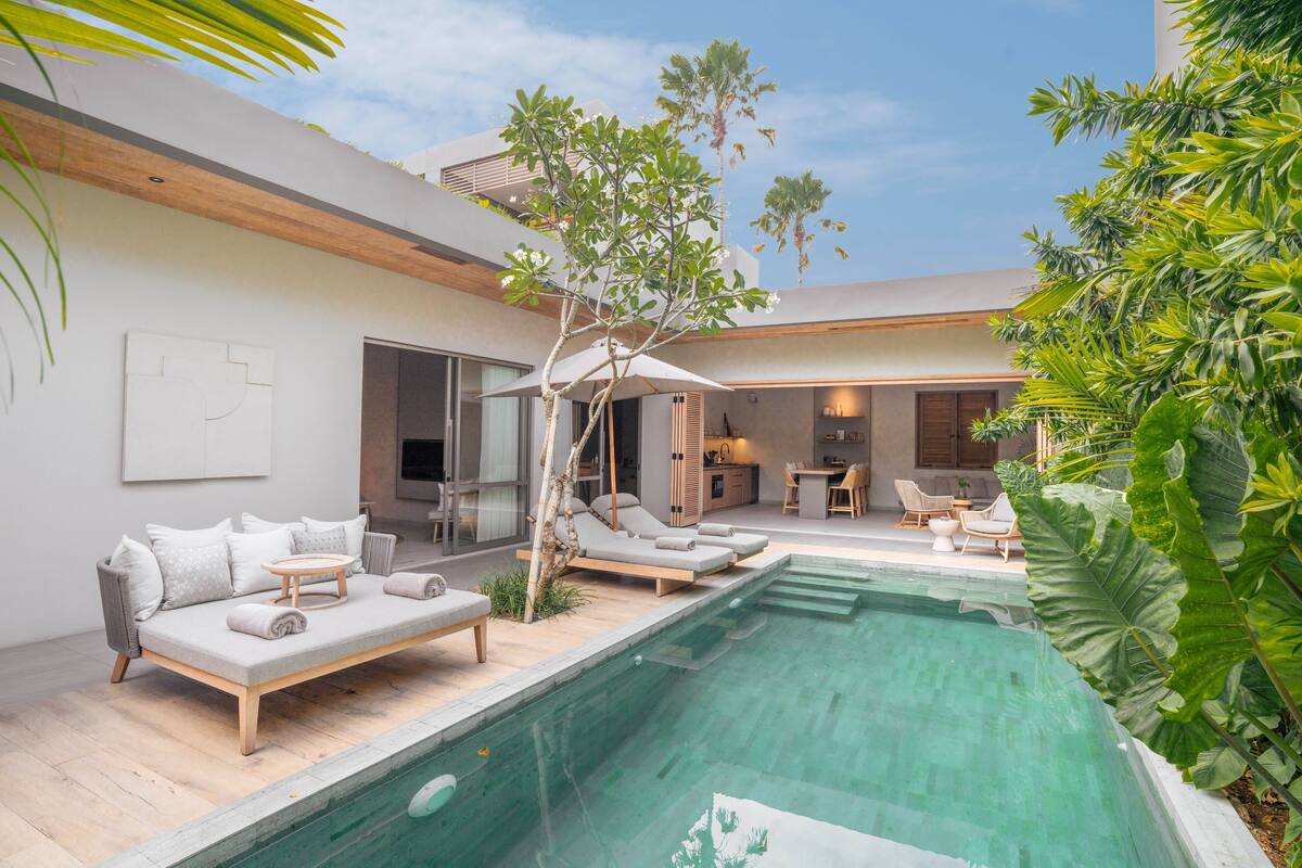 A modern outdoor pool area with a built-in sofa and lounge chairs, surrounded by lush greenery and a clear sky.
