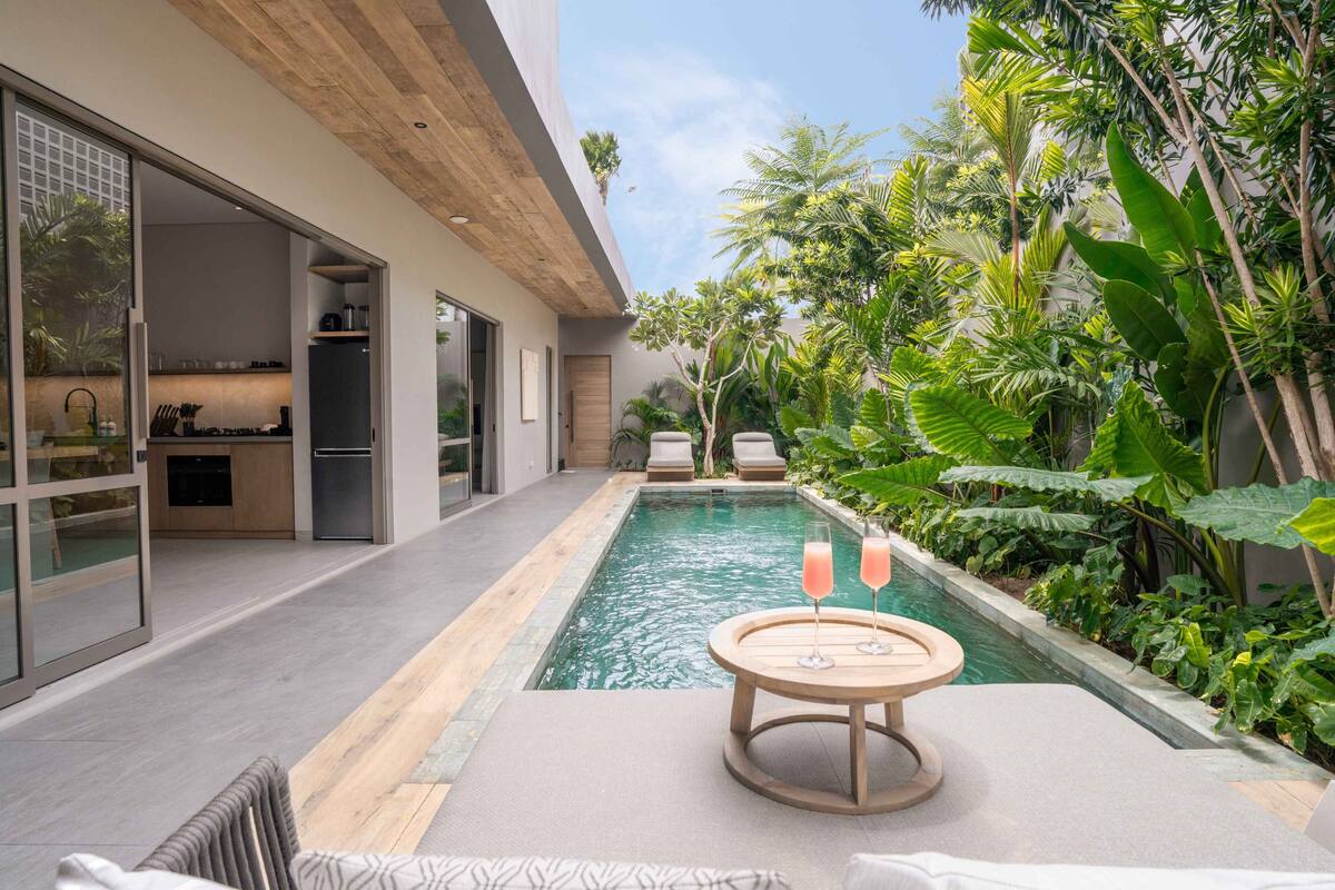 A modern outdoor pool area with a wooden table and lounge chairs, surrounded by lush tropical plants.
