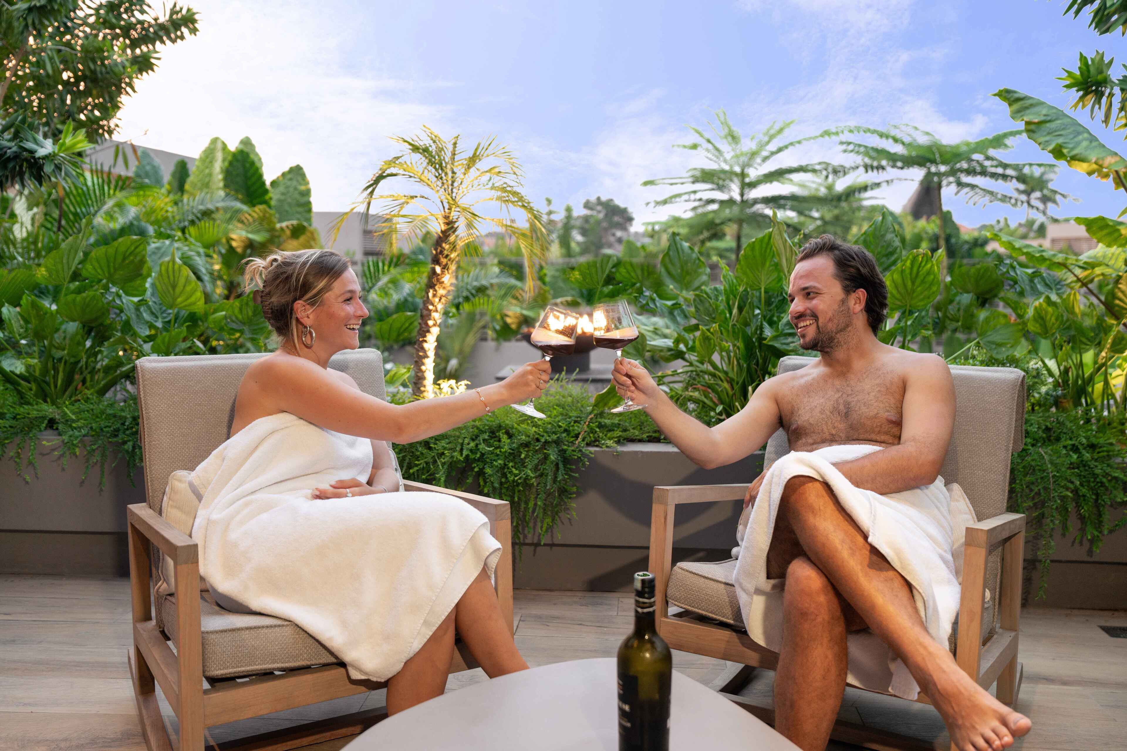 Two people seated on cushioned chairs outdoors, holding glasses with a drink, with a bottle on the table and lush greenery in the background.