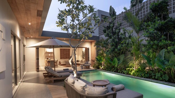 Un área de piscina moderna al aire libre con un sillón de descanso y sombrilla, rodeada de exuberante vegetación y una terraza de madera.