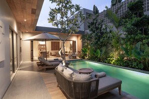 Un área de piscina moderna al aire libre con un sillón de descanso y sombrilla, rodeada de exuberante vegetación y una terraza de madera.