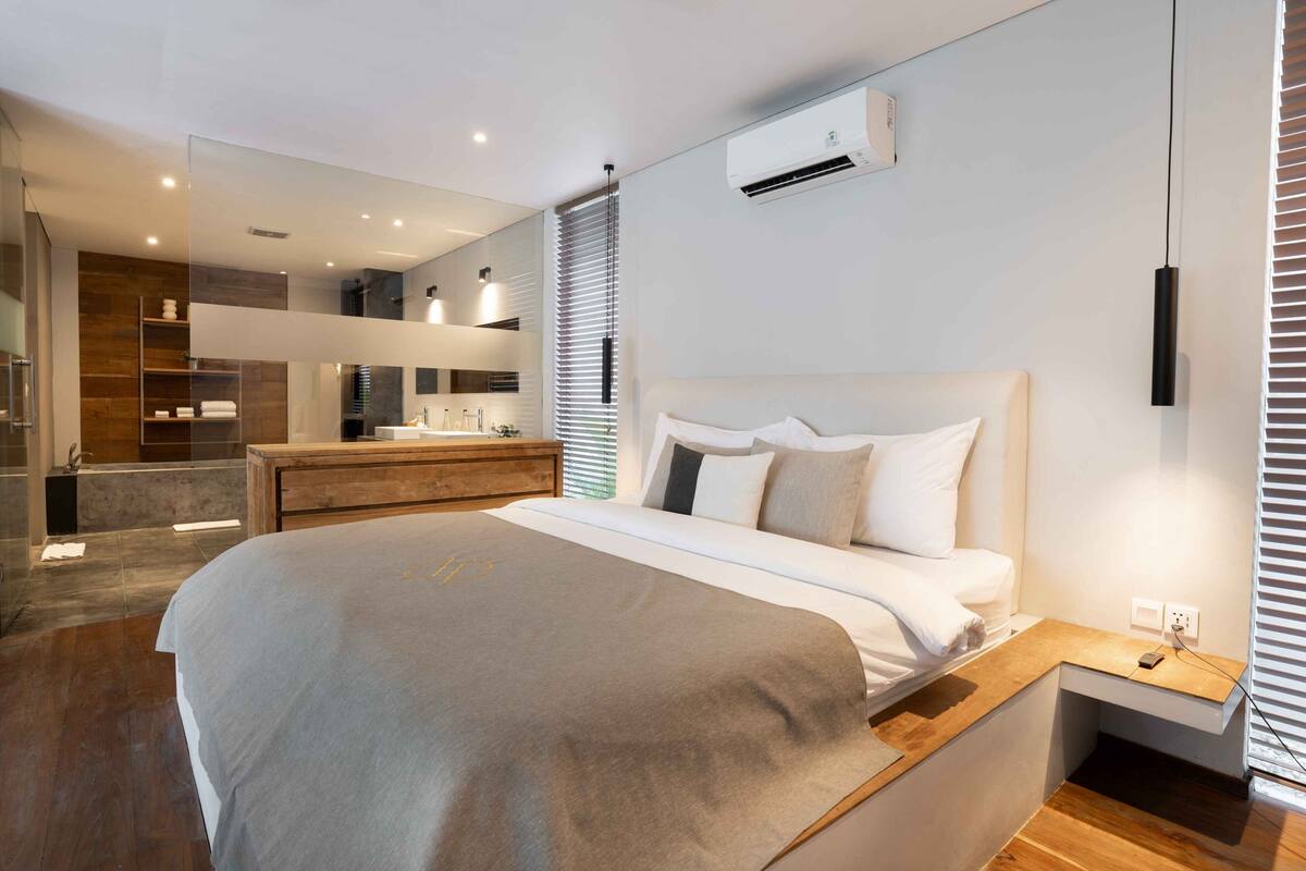 A modern bedroom with a large bed, bedside tables, a built-in bench, a wall-mounted air conditioner, and a bathroom visible through a glass door.