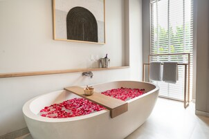 Ein modernes Badezimmer mit einer freistehenden Badewanne, Rosenblättern, einem Holzregal, einer Schale und einem gerahmten Kunstwerk an der Wand.