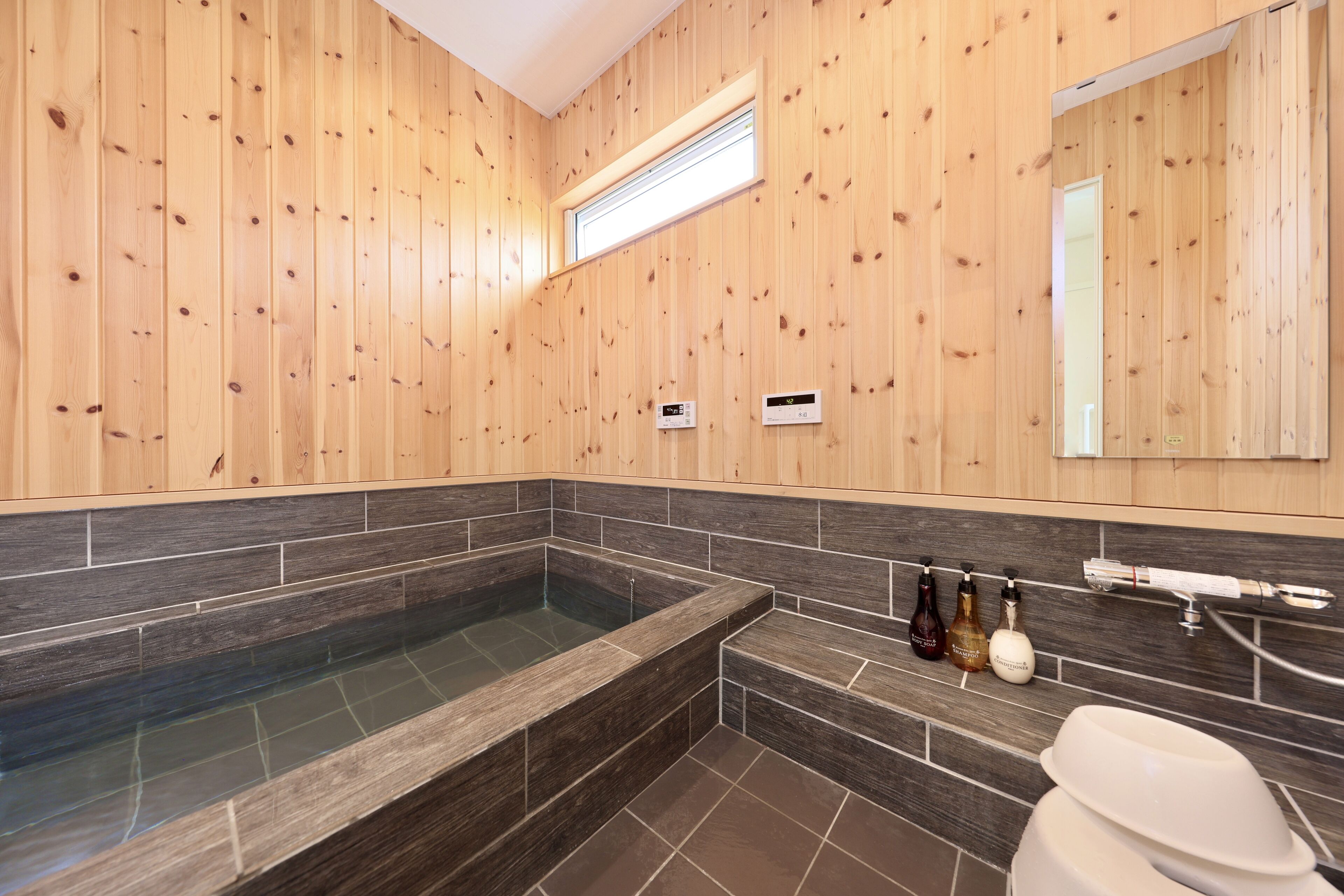 A modern bathroom with wooden walls, a large bathtub, dark tiled flooring, and a built-in sink with toiletries.