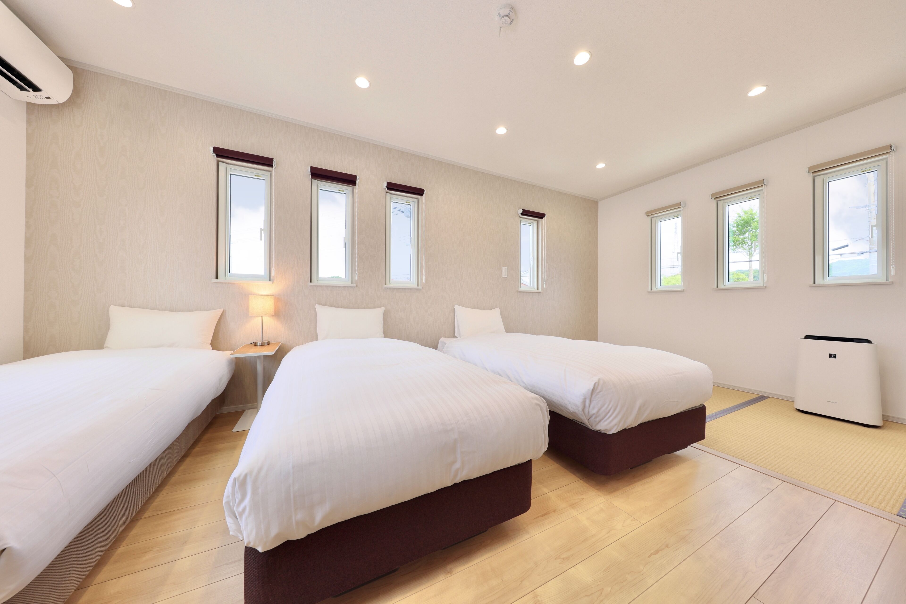 A hotel room with two single beds, a bedside table with a lamp, a heater, and large windows with blinds.