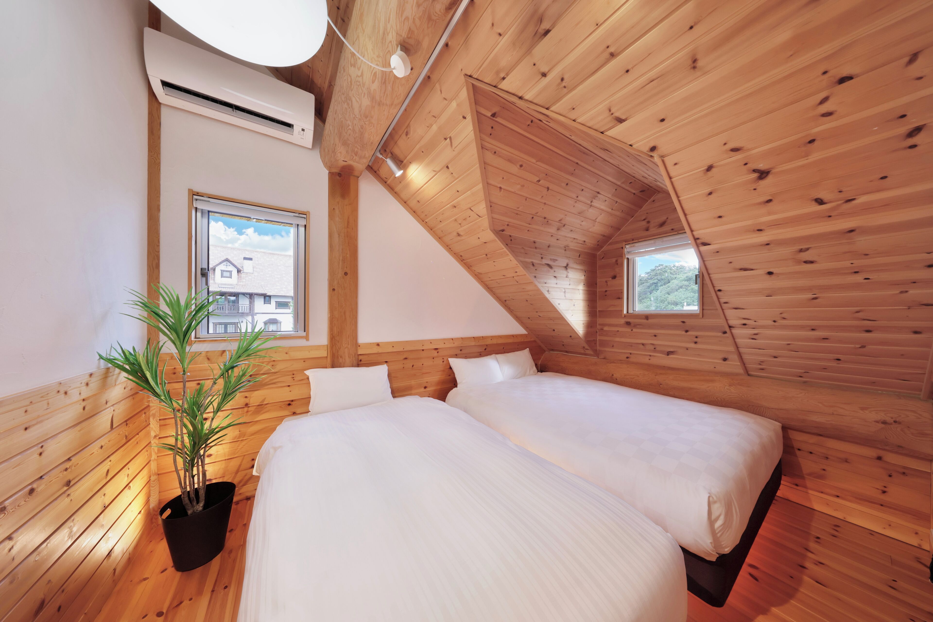 Camera da letto con due letti, pareti in legno, una pianta in vaso e una finestra con vista su una casa.