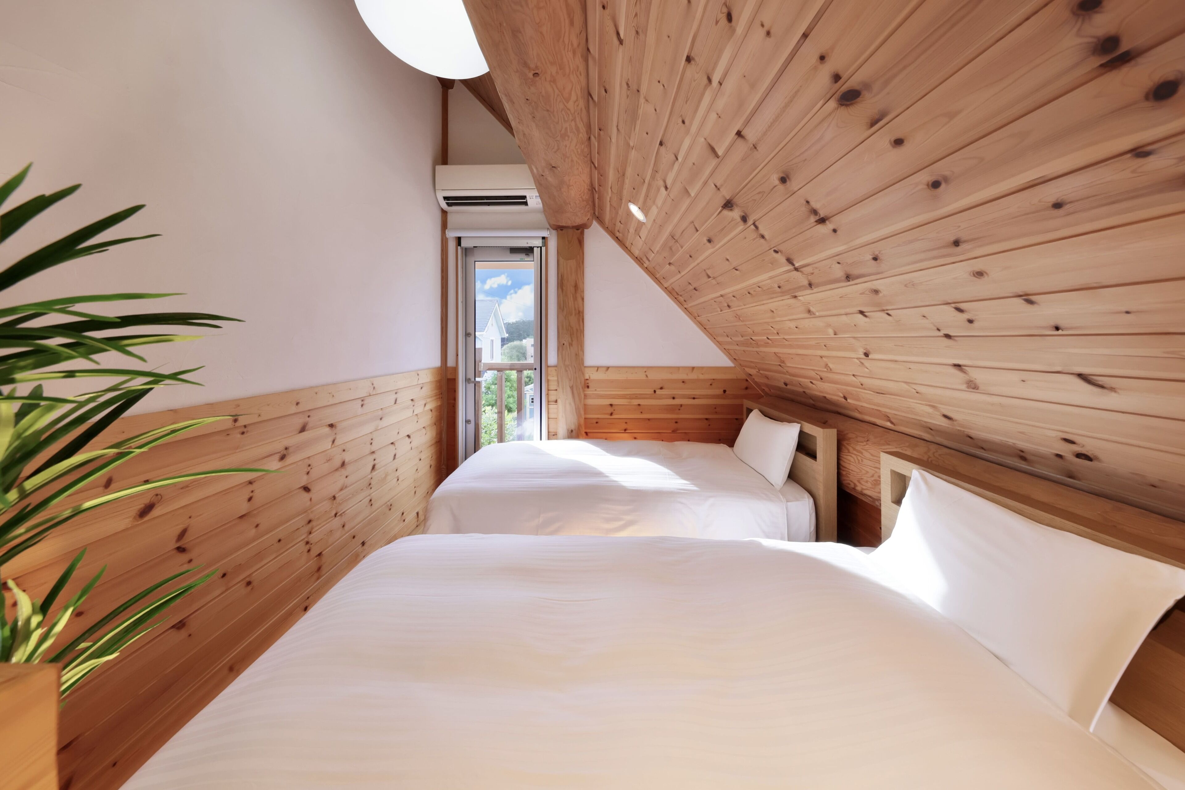 A room with wooden walls and a sloped ceiling, two beds with white linens, a potted plant, and a window with a view of greenery.