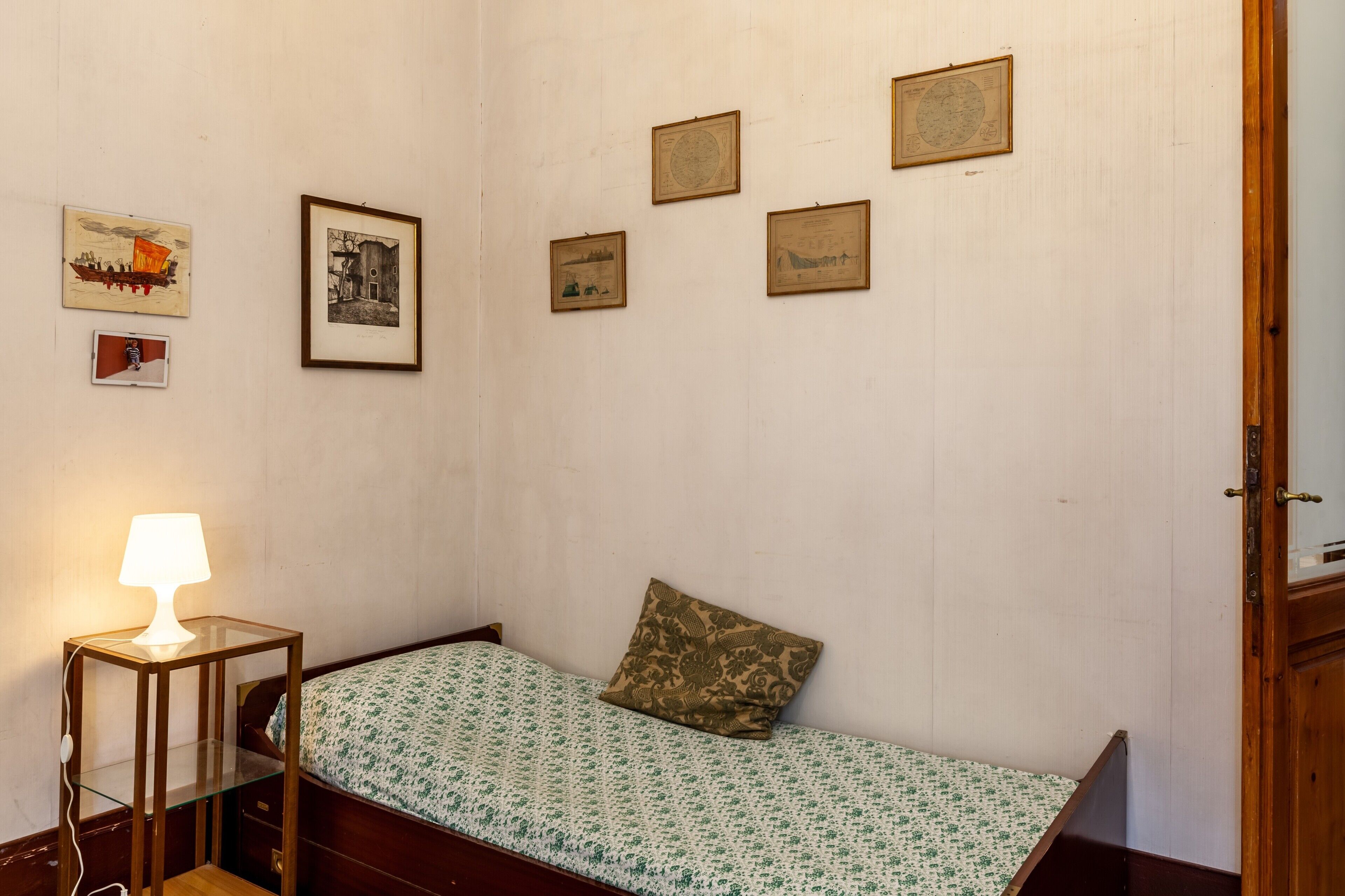 Ein kleines Zimmer mit einem Einzelbett, einem Nachttisch mit Lampe und mehreren gerahmten Bildern an der Wand.