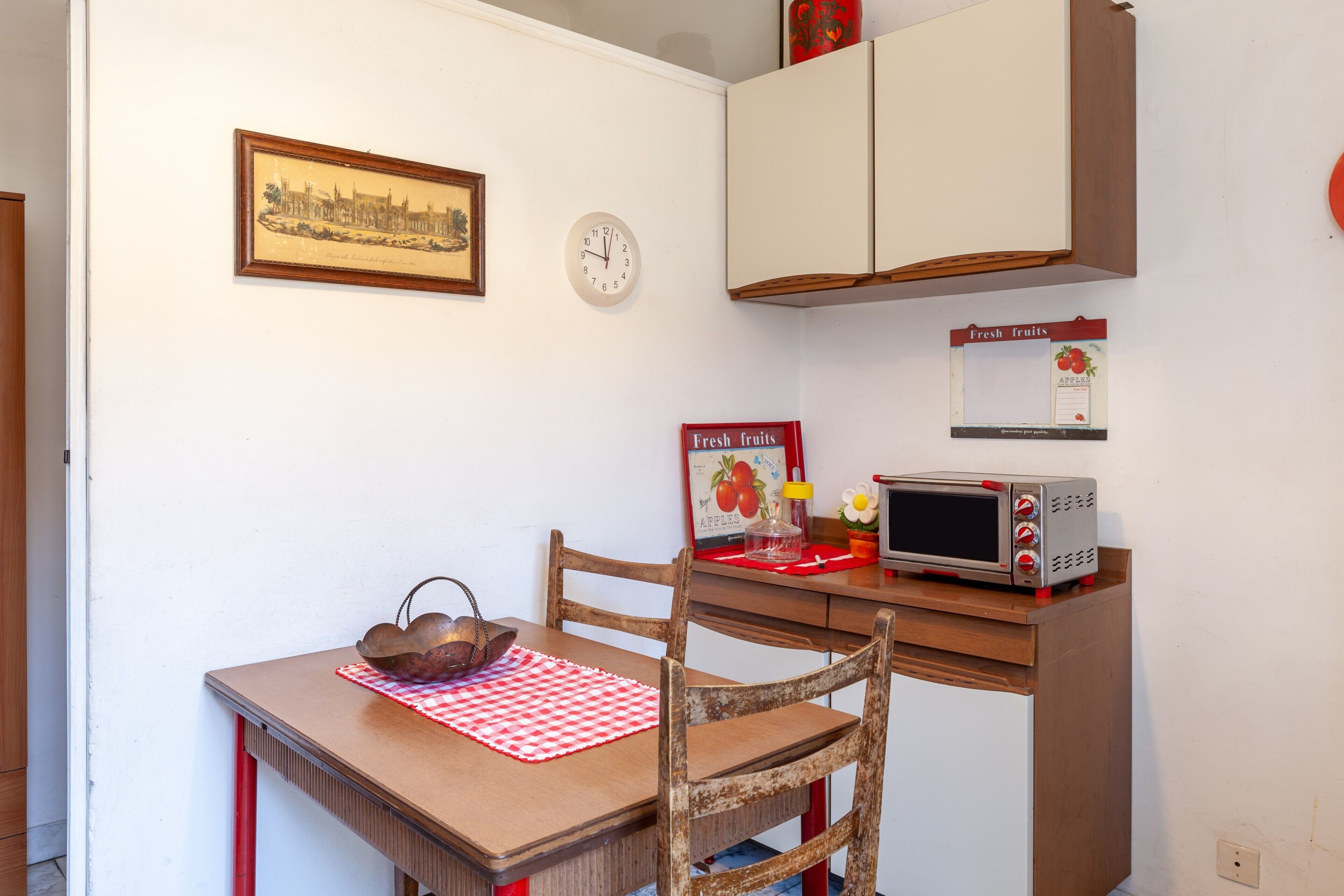 A kitchen with a wooden table, chairs, a microwave, a clock, and a framed picture on the wall.
