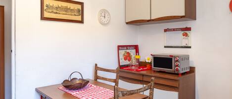 A kitchen with a wooden table, chairs, a microwave, a clock, and a framed picture on the wall.