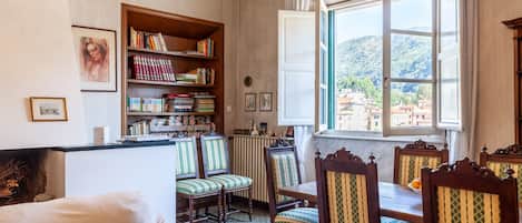 A room with a dining table, chairs, a bookshelf, a radiator, and a window with a view of buildings.