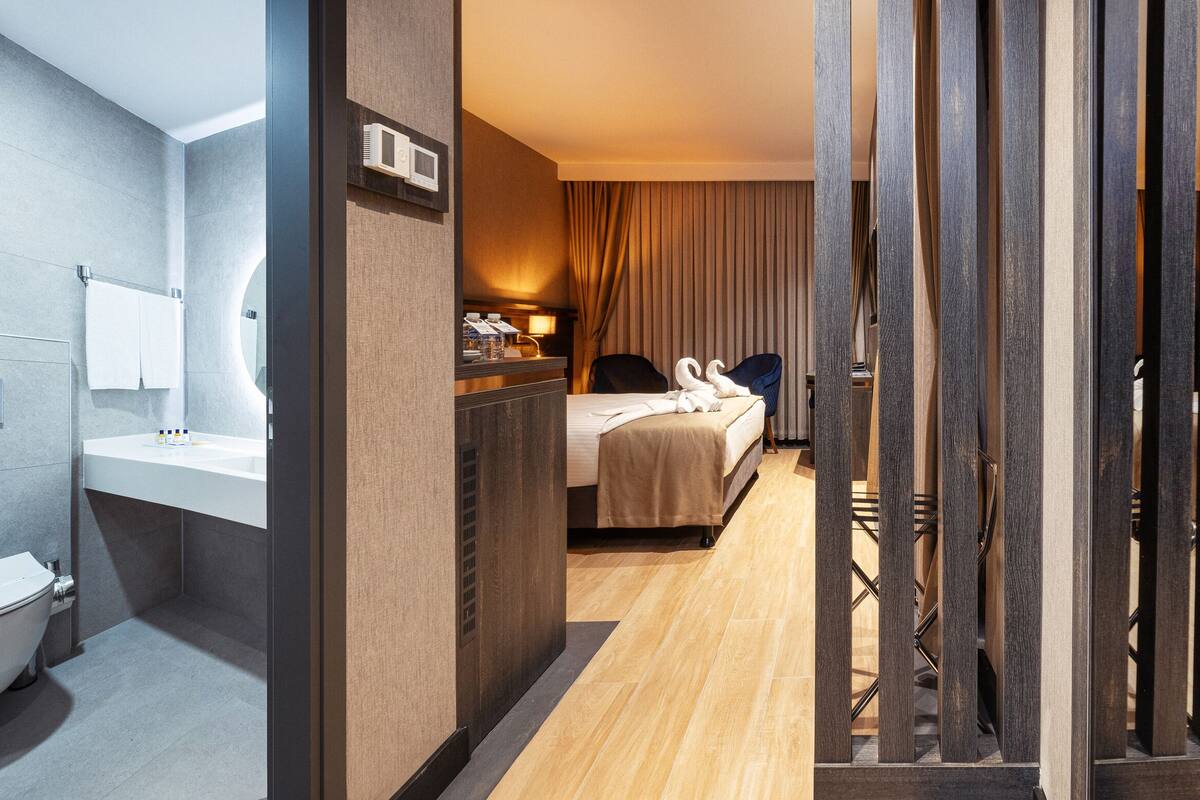 Een moderne hotelkamer met een houten vloer, een bed met een beige deken, een nachtkastje met een lamp en een grote spiegel.