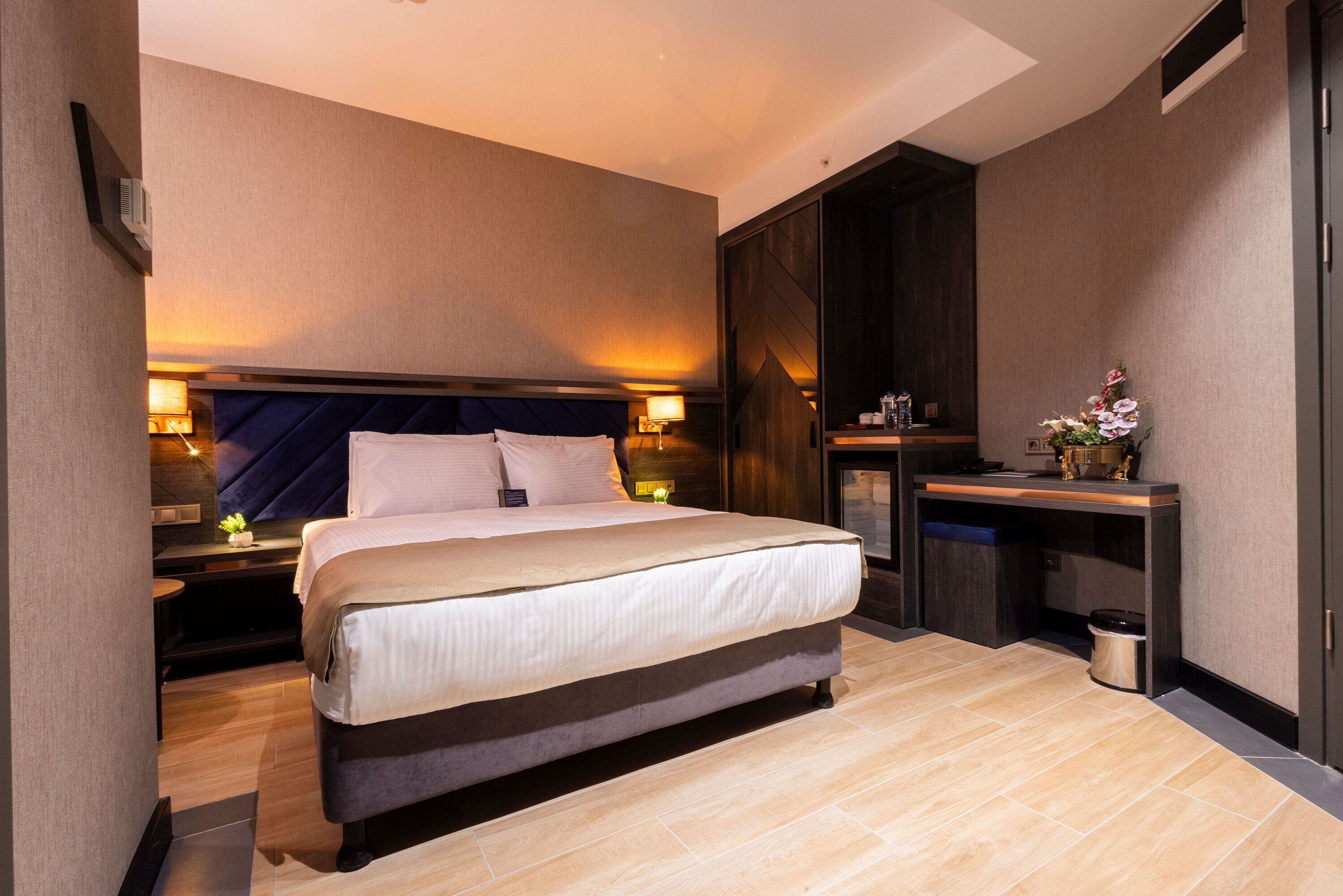A modern hotel room with a large bed, bedside tables, a desk, and a small trash bin.