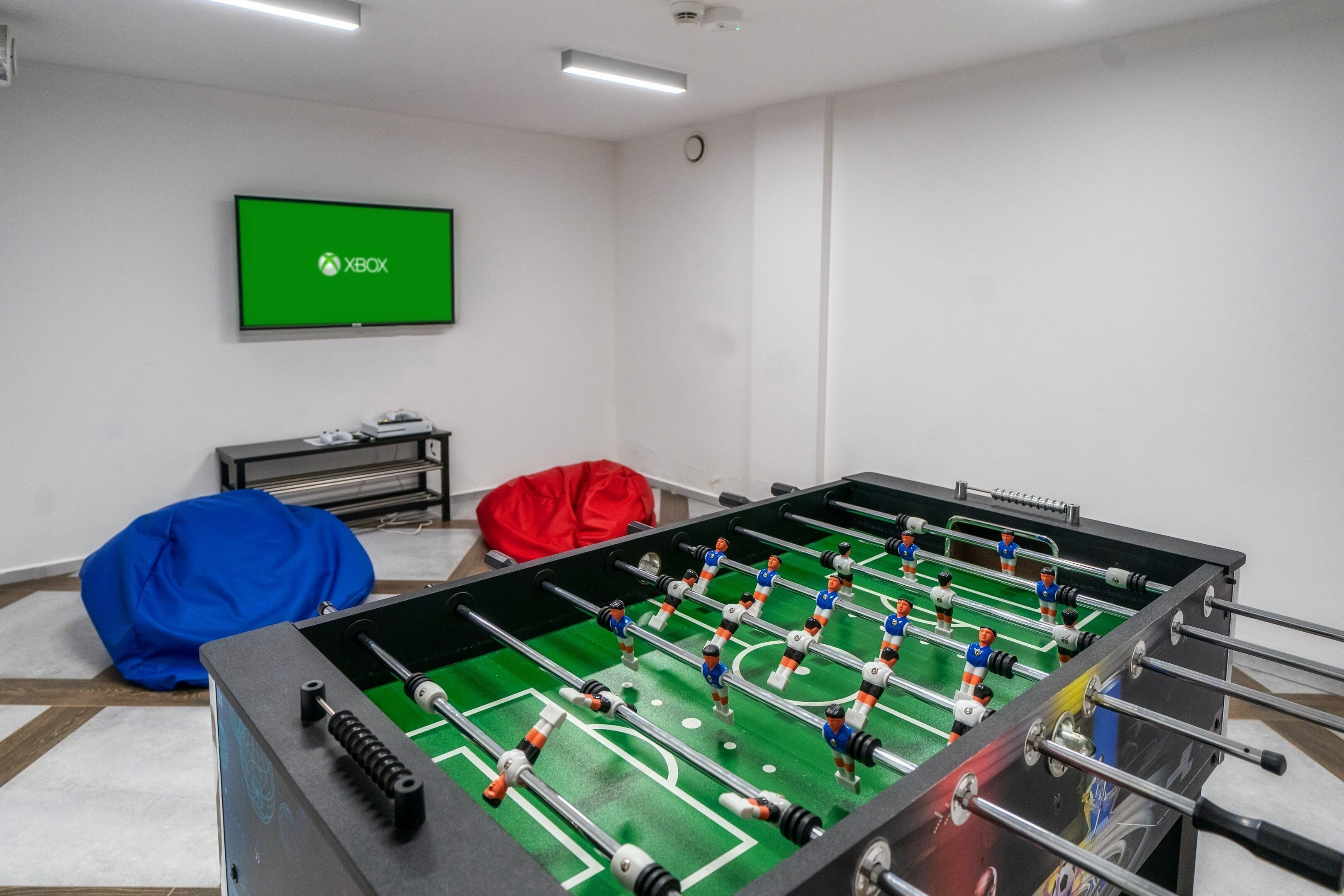 Una habitación con mesa de futbolín, televisor mostrando el logo de Xbox y un sillón de peluche azul.