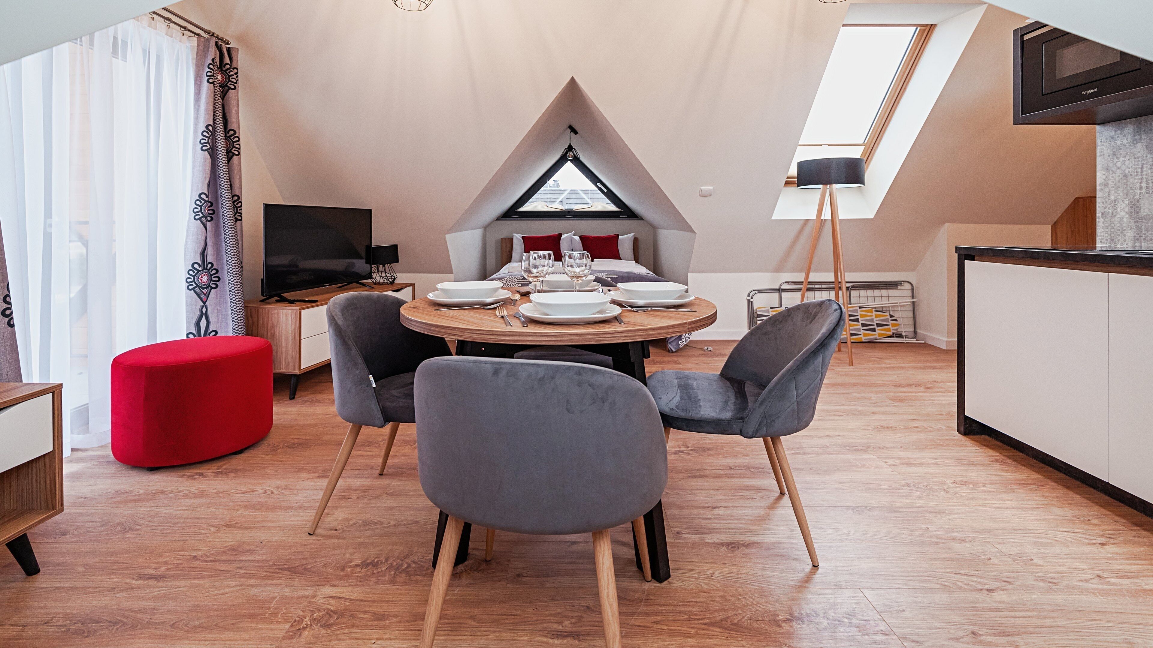 A modern dining area with a round table, grey upholstered chairs, and a red ottoman. A flat-screen TV is mounted on the wall, and a window with a black frame is visible.
