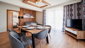 Uma sala de jantar moderna com uma mesa de madeira posta para quatro, cadeiras cinzentas estofadas, uma TV de tela plana em um suporte de madeira e um quadro na parede.