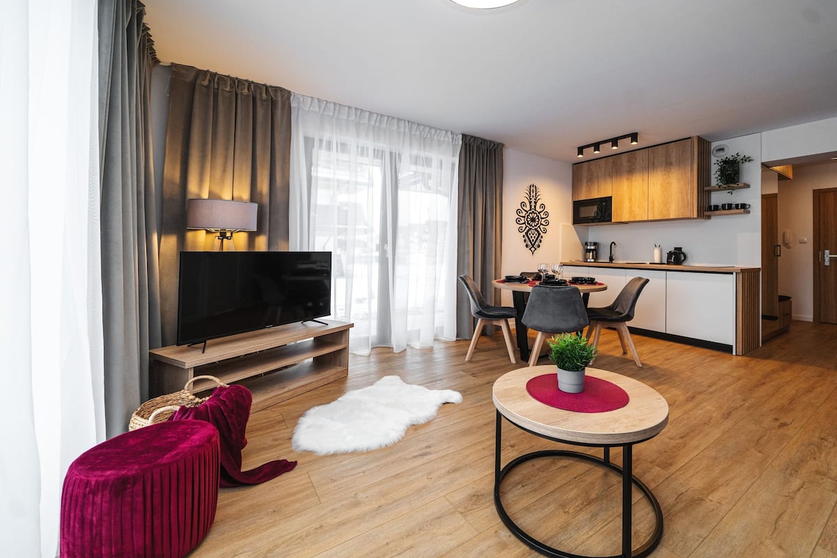 Ein modernes Wohnzimmer mit Flachbildfernseher, einem roten Hocker und einem Essbereich mit rundem Tisch.