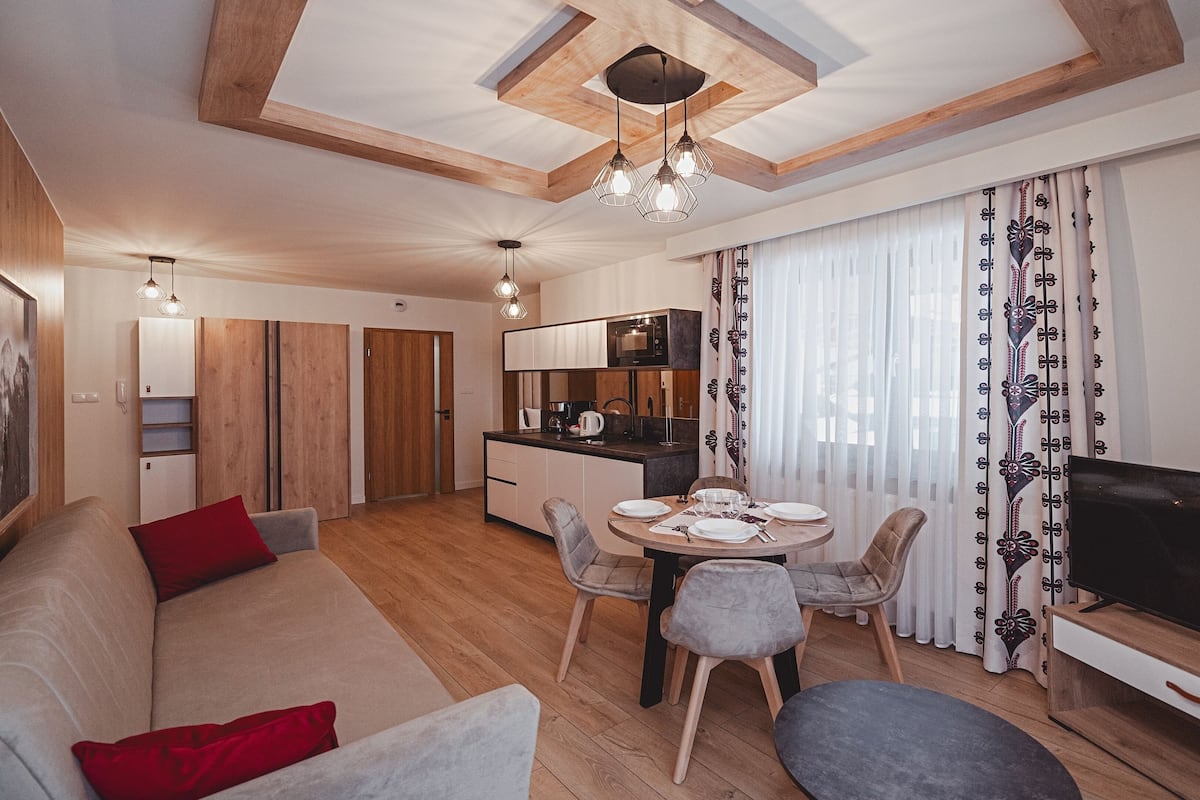 Ein modernes Wohnzimmer mit Sofa, Esstisch und Stühlen. Es gibt eine Küchenzeile mit Mikrowelle und Fernseher. Der Raum hat Holzböden und eine Decke mit sichtbaren Balken.