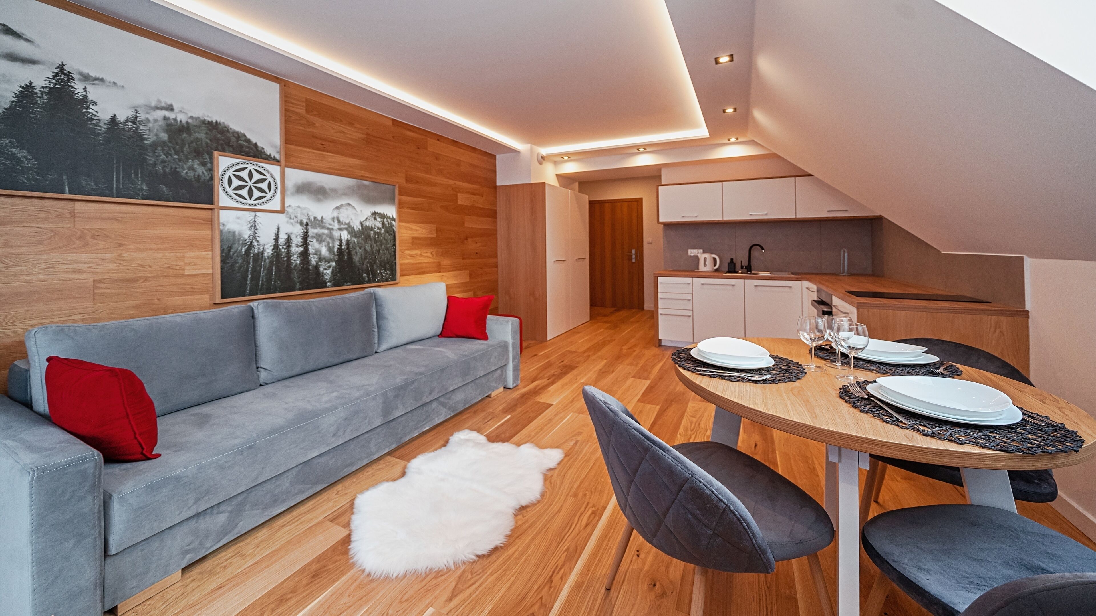 A modern living room with a grey sofa, a round wooden dining table set with plates and glasses, and a white furry rug on the wooden floor.