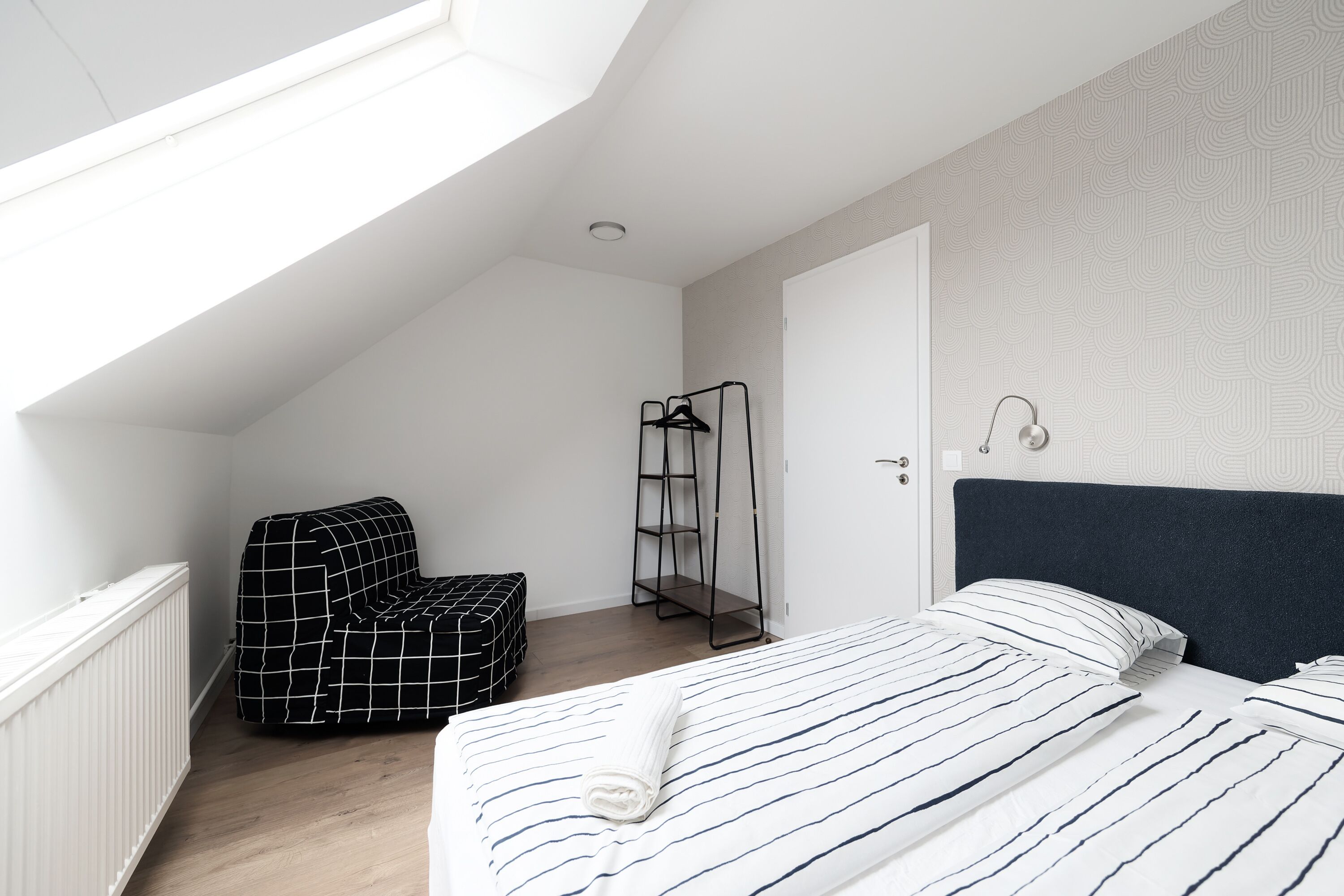 Ein Schlafzimmer mit schrägverdeckter Decke, einem Bett mit gestreifter Bettwäsche, einem schwarz-weiß karierten Stuhl und einem Metallregal.