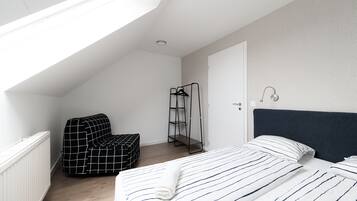 Ein Schlafzimmer mit schrägverdeckter Decke, einem Bett mit gestreifter Bettwäsche, einem schwarz-weiß karierten Stuhl und einem Metallregal.