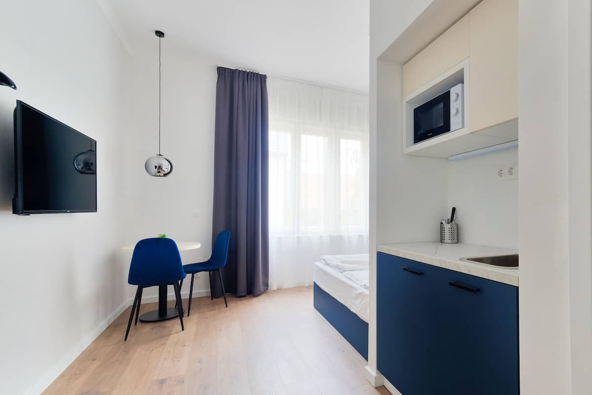 A modern bedroom with a bed, a desk, a chair, a TV, and a kitchenette with a microwave.