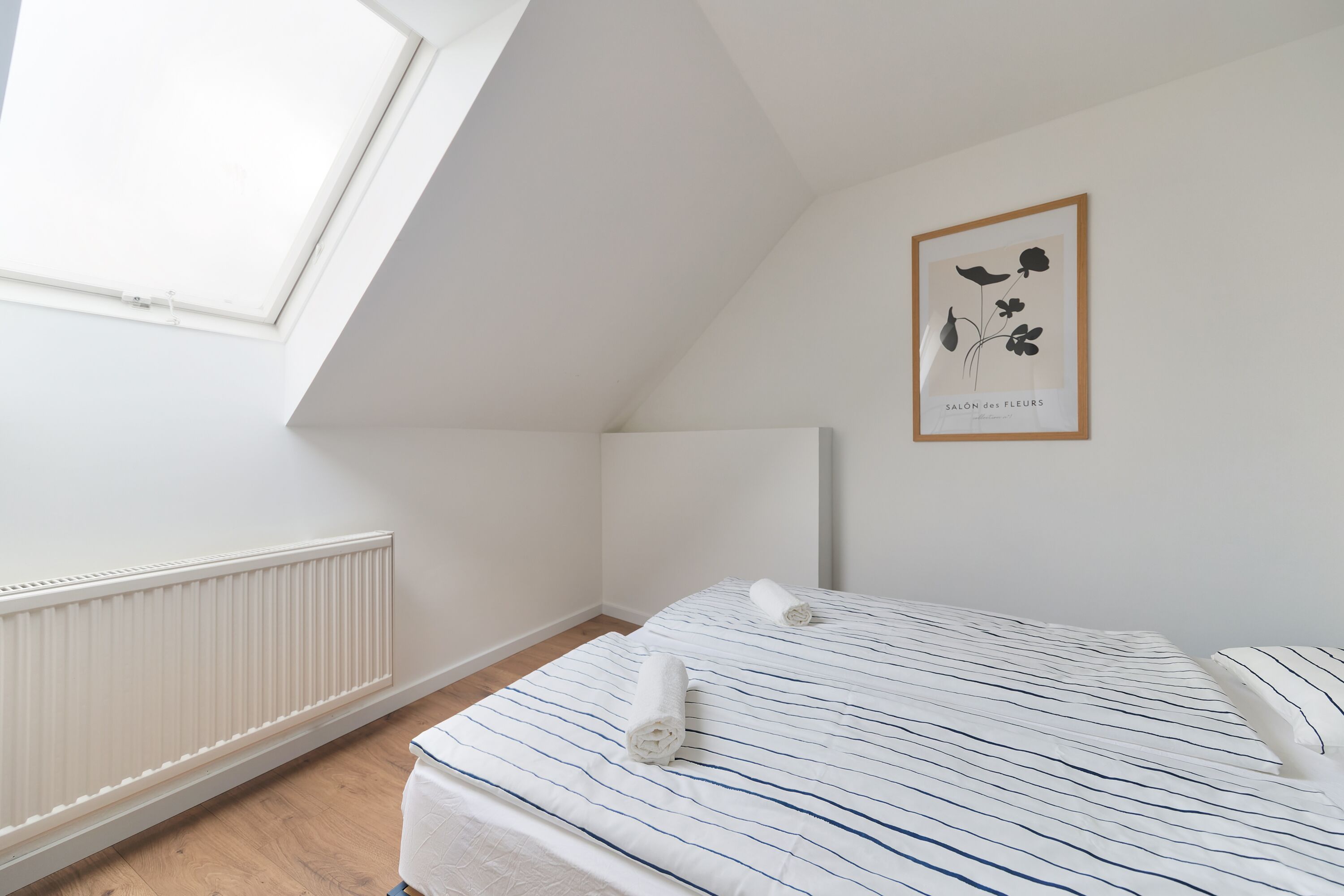 Une chambre avec un plafond en pente, un puits de lumière, un radiateur, un lit avec une literie à rayures et une œuvre d’art florale encadrée accrochée au mur.