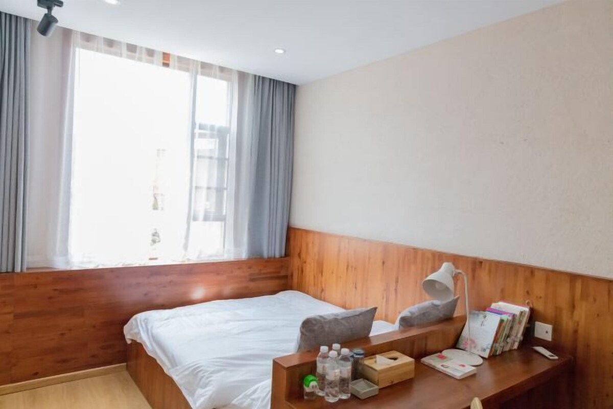 Hotelkamer met houten hoofdeinde, een bed met wit beddengoed, nachtkastje met lamp, raam met gordijnen en uitzicht op gebouwen.