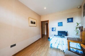 Une pièce avec un plancher en bois, un lit recouvert d’une couverture bleue et blanche, un bureau avec un ordinateur et une chaise.
