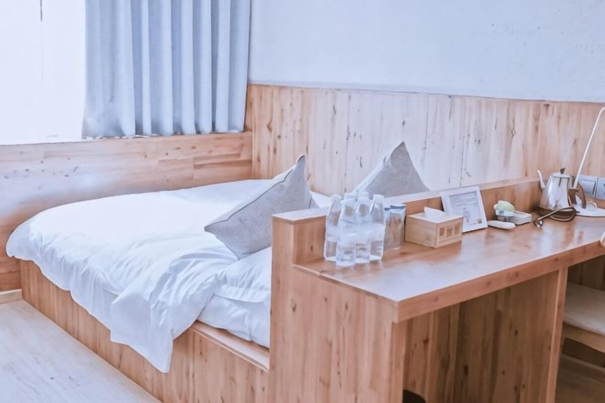Een houten bed met wit beddengoed, een nachtkastje van hout met een lamp, en een raam met gordijnen.