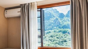 Una habitación con cama, aire acondicionado, ventana con cortinas y vista a una montaña boscosa.