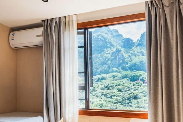 A room with a bed, an air conditioning unit, a window with curtains, and a view of a forested mountain. - Manman Qingfeng Hostel (Xingyi)