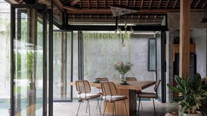 A modern dining area with a wooden table and chairs, large windows, and hanging plants. - Pritidaya Private Pool Villa Canggu by OXO (Canggu)