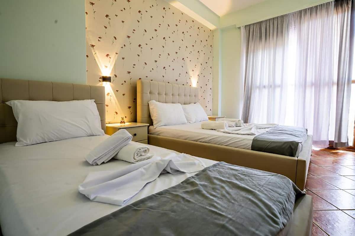 A hotel room with two beds, a floral wallpaper, a nightstand with a lamp, and a window with curtains.