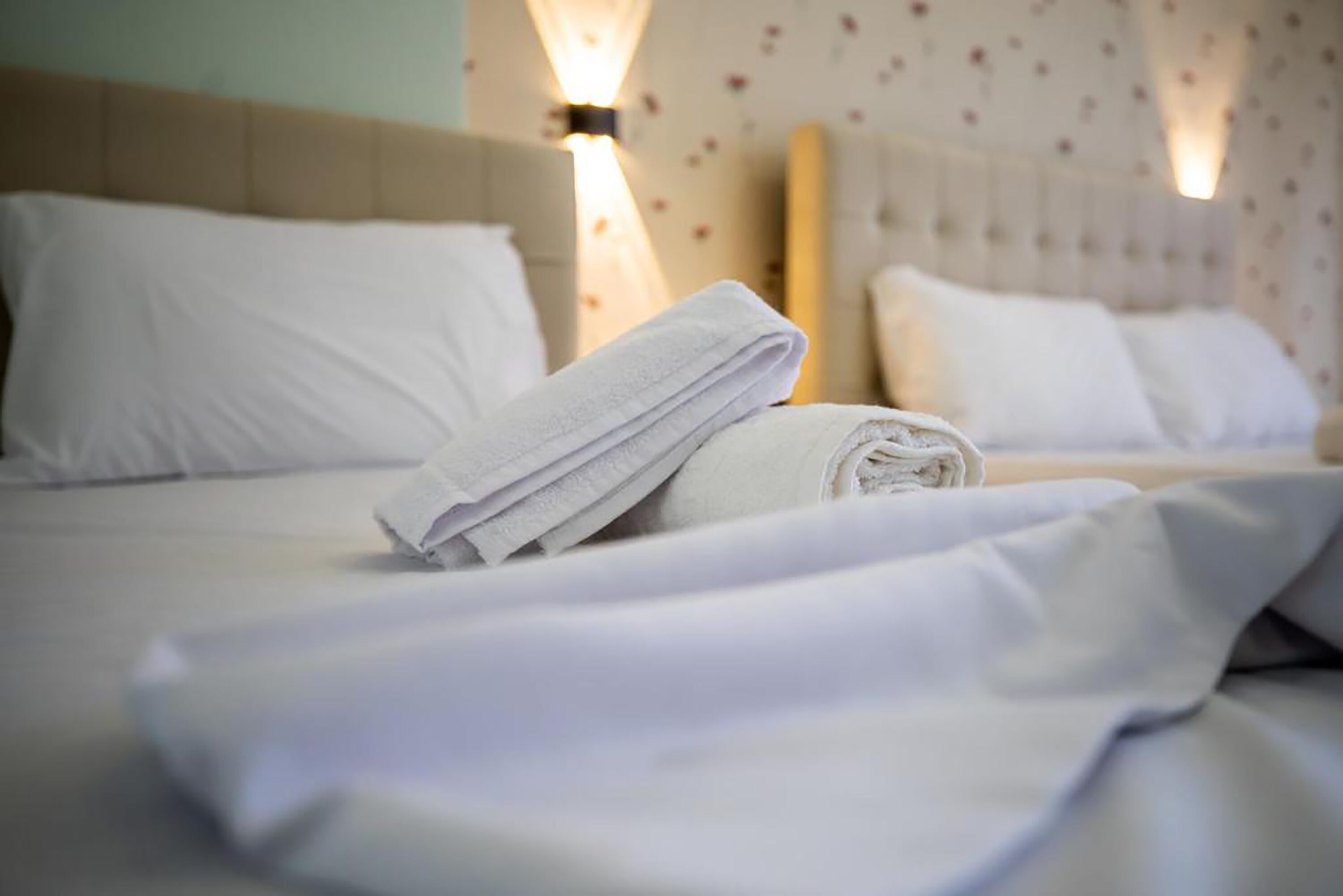 Una cama bien tendida con sábanas blancas y una toalla enrollada encima, en una habitación con estampado floral en el papel tapiz y un cabecero de color claro.