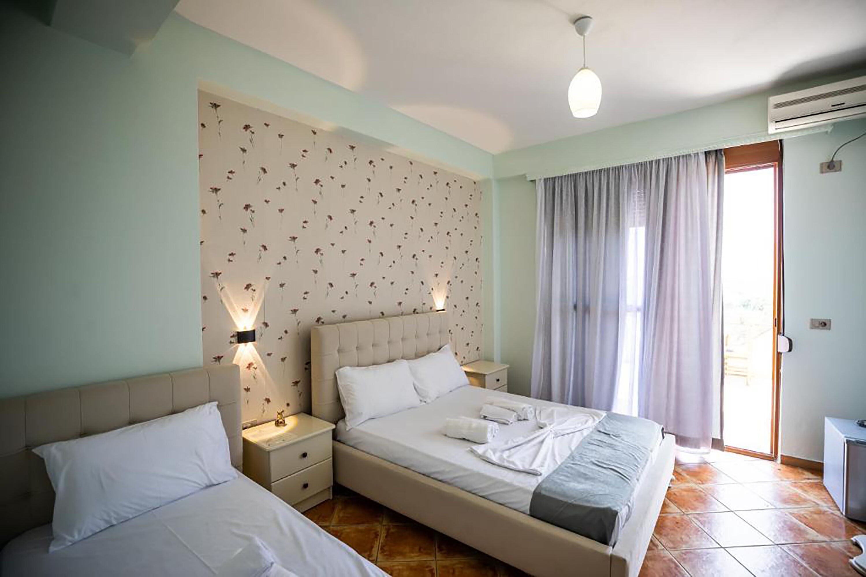 A double bed with a headboard, bedside tables, a wall-mounted light, and a window with curtains.