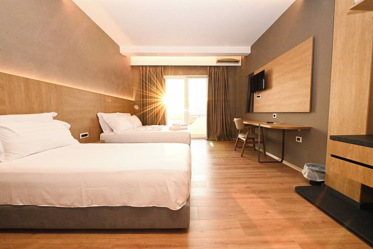 A hotel room with two beds, a desk, a TV, and a large window with curtains.