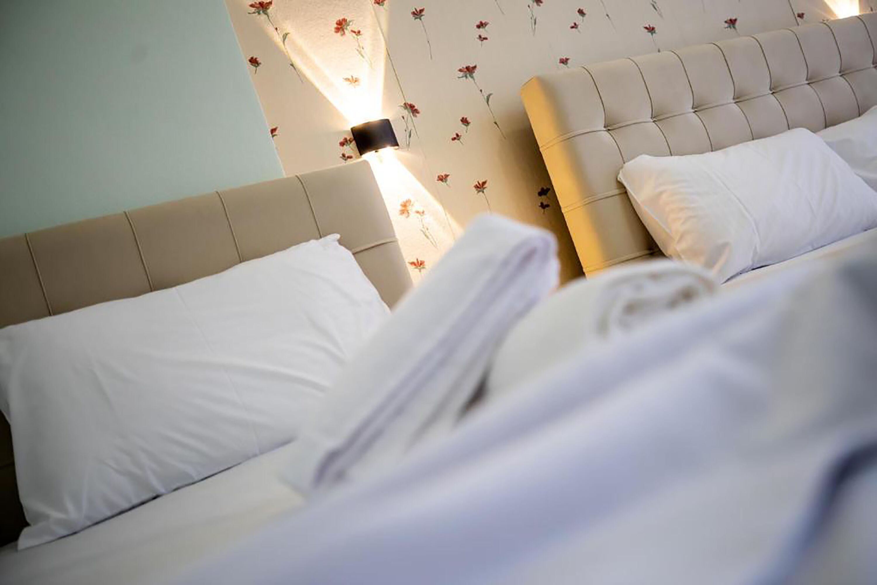 A neatly made bed with white linens and pillows, a tufted headboard, and a floral wallpaper pattern.